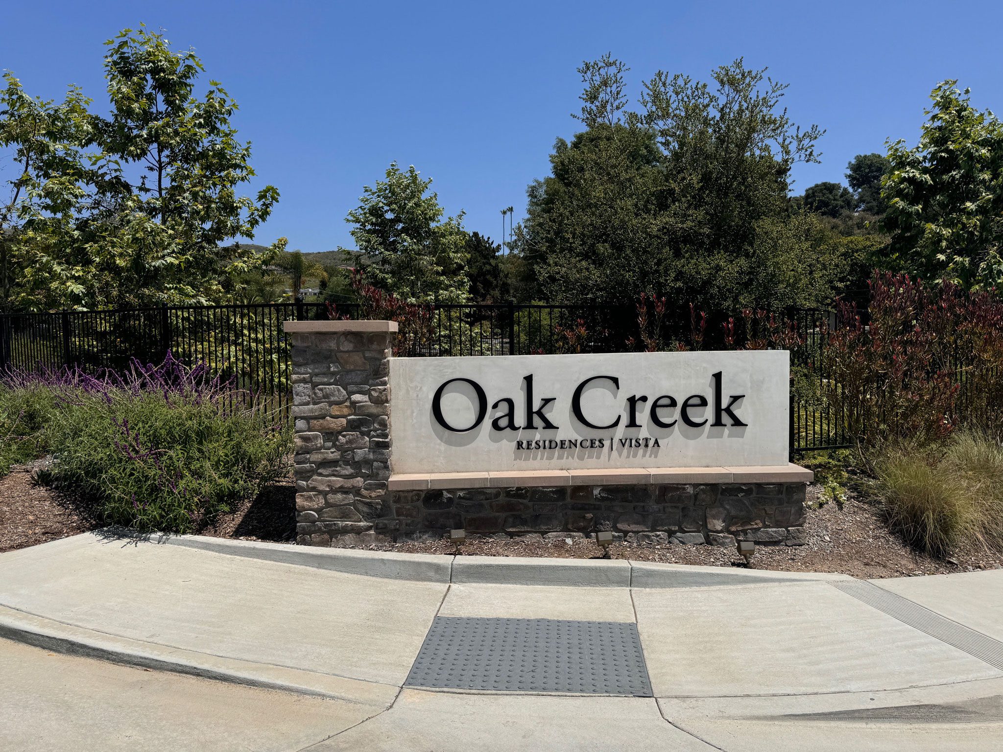IMG_2711 A sign with "Oak Creek Residences Vista" inscribed on it, situated next to a stone pillar and surrounded by greenery and a black fence under a clear blue sky.