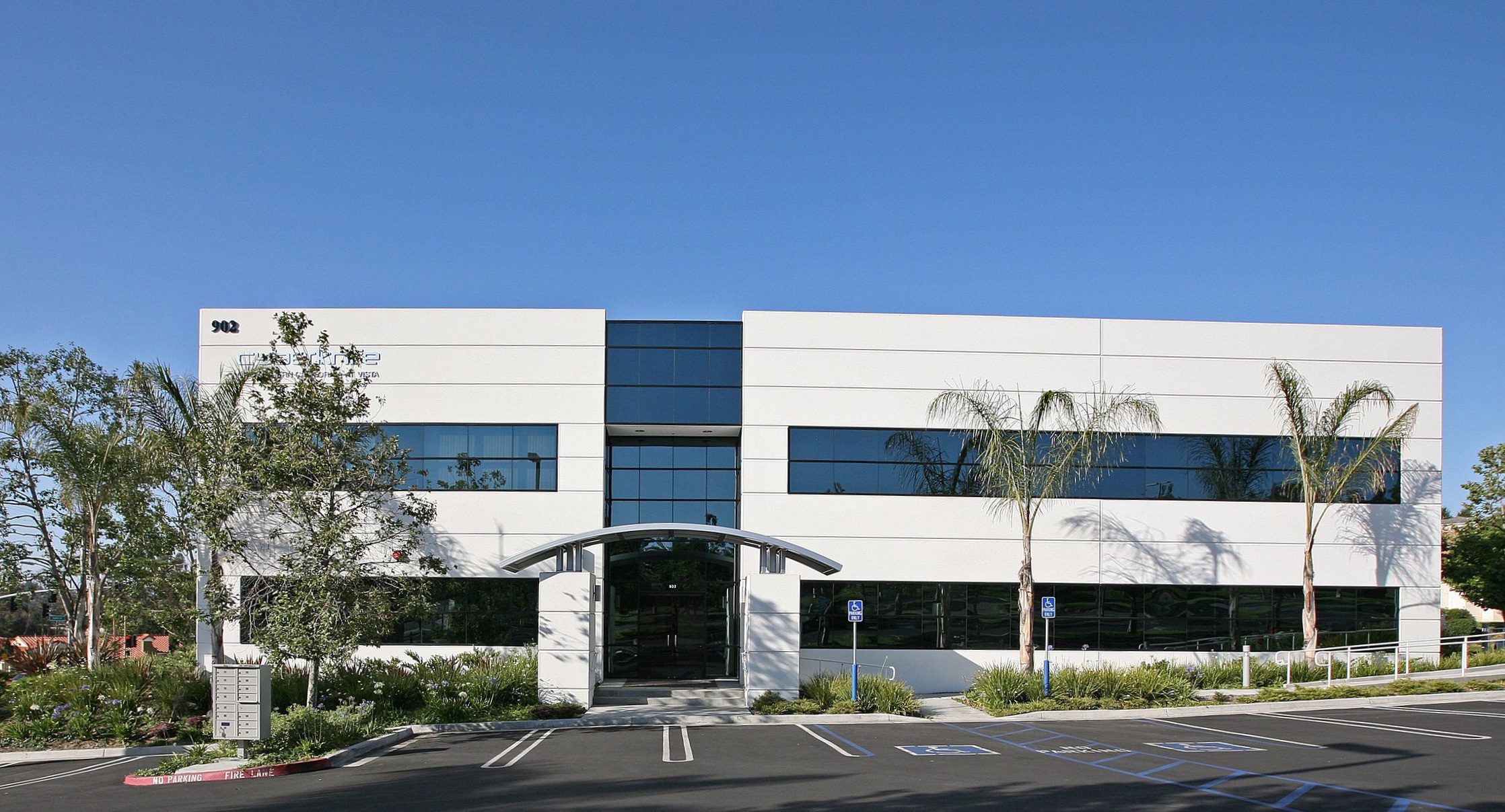 IMG_3714 Modern two-story office building with large reflective windows and an arched entrance, surrounded by palm trees and a parking lot with marked parking spaces.