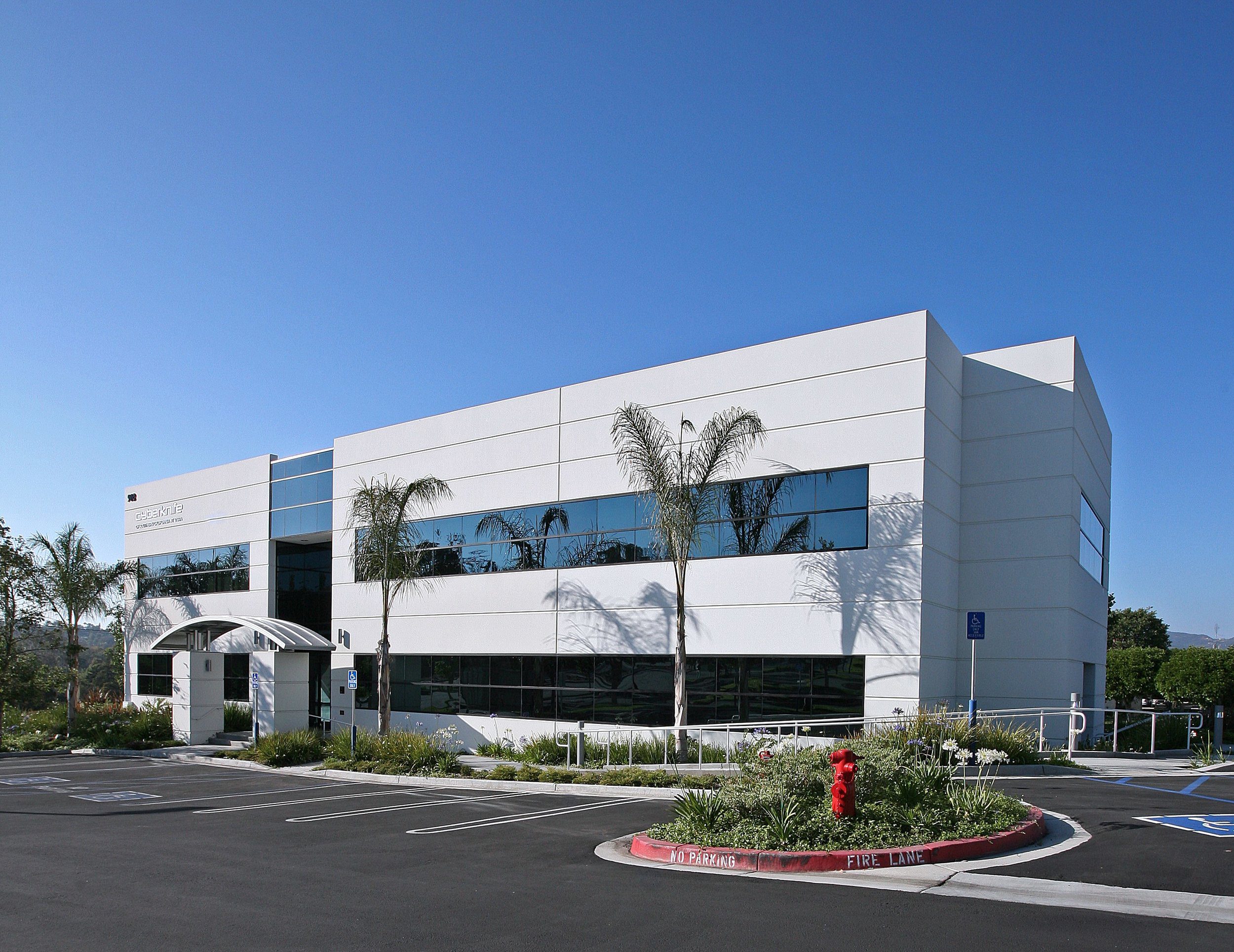 IMG_3731 A modern two-story office building with large windows, palm trees in front, and a landscaped parking area under a clear blue sky.