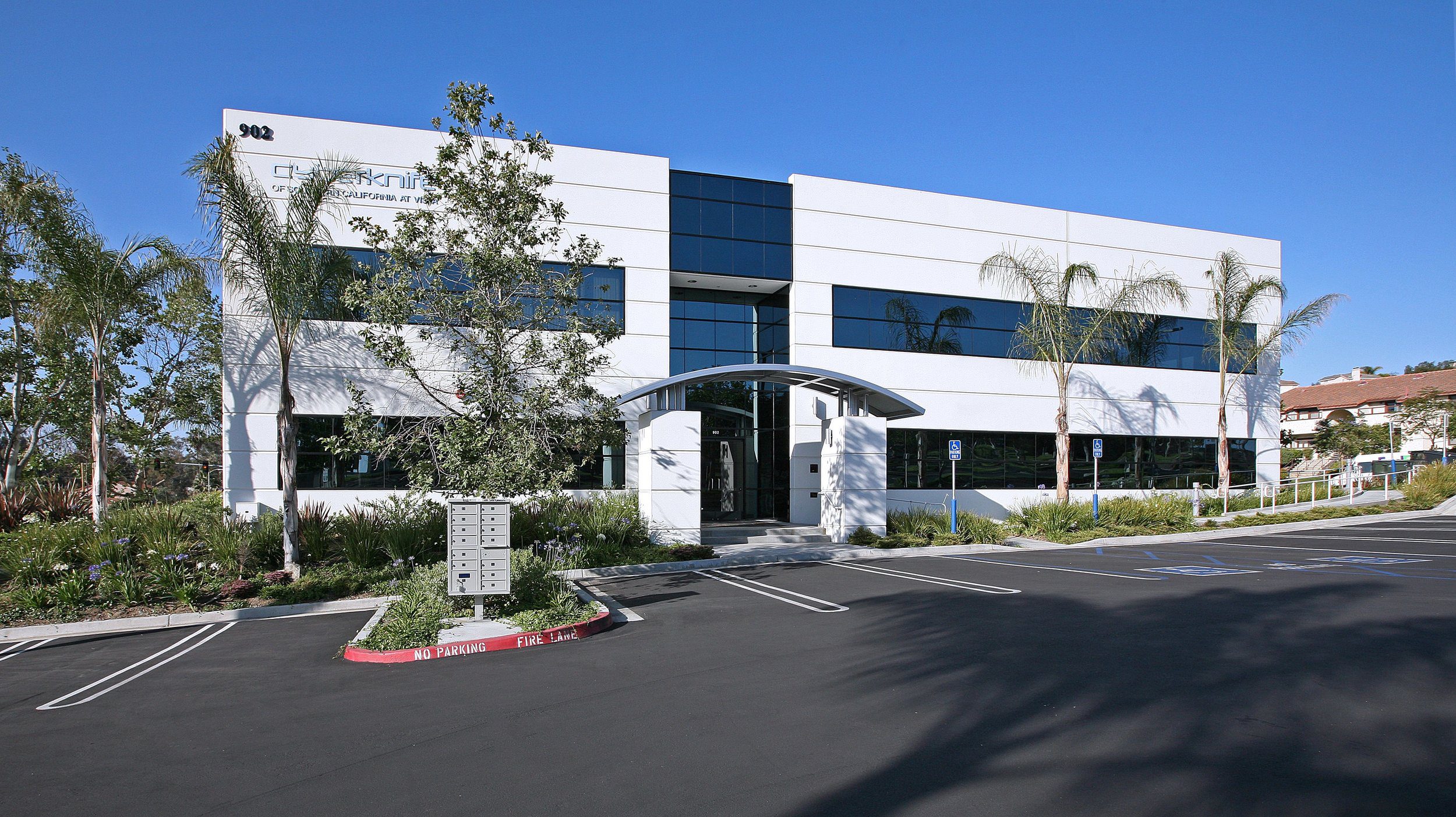 IMG_3734 A modern, two-story commercial building with large windows and an arched entrance, surrounded by palm trees and landscaping, with an adjacent parking lot under a clear blue sky.